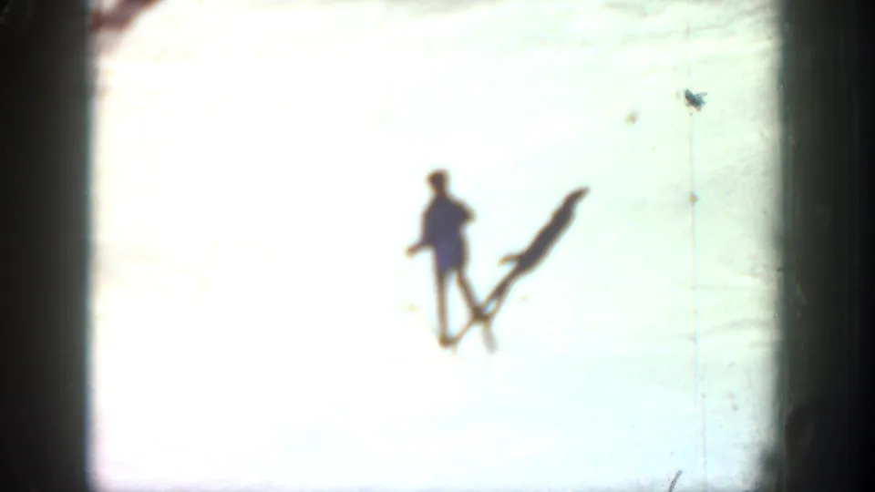 A skier is seen on a bright overexposed snowfield, with a long shadow and dark film edge visible in the 8mm frame.
