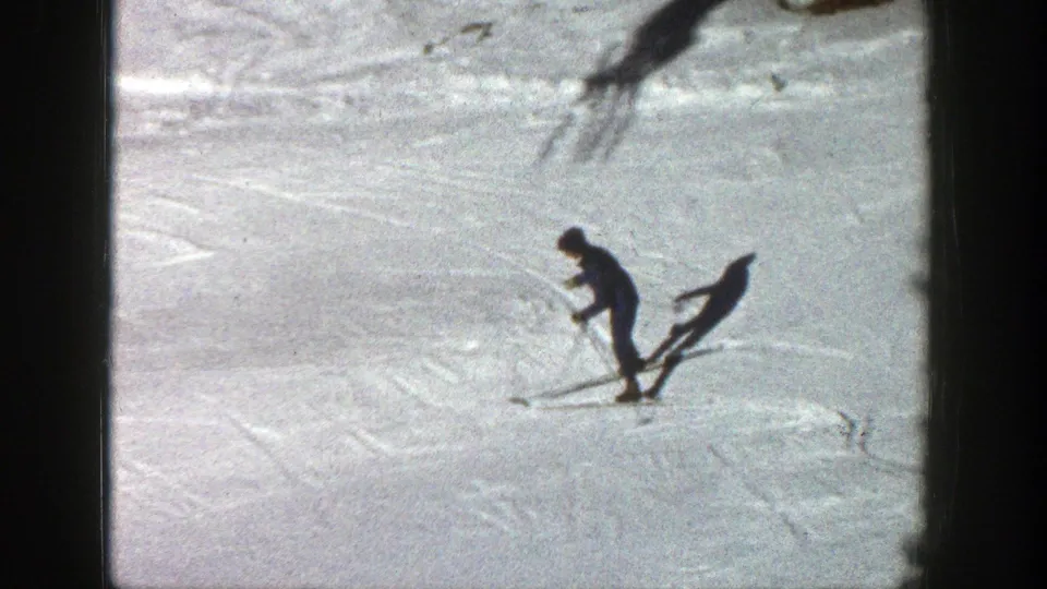 An adult skier moves across the Aspen snow, casting a long dark shadow on the textured slope in grainy 8mm footage.