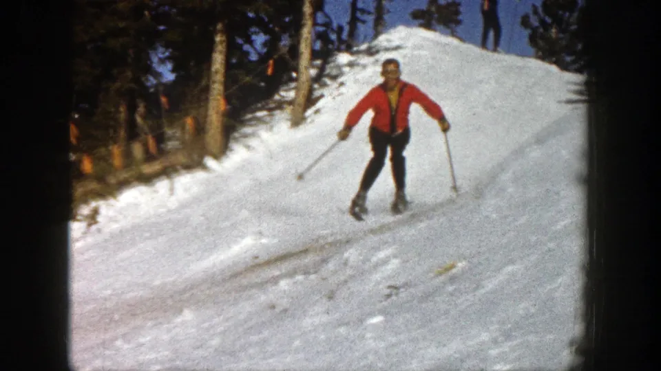 Adult skier in a bright red jacket and dark pants descends a tree-lined Aspen ski slope with poles.