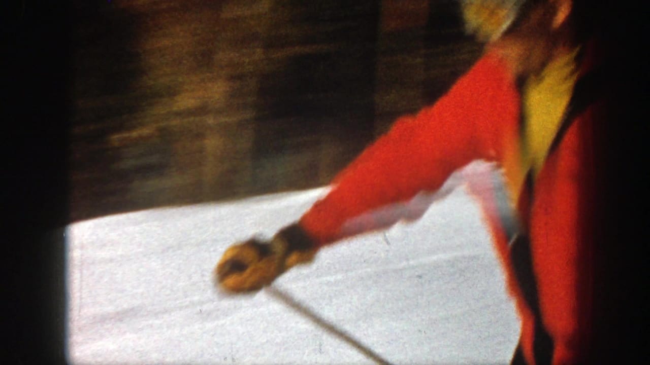 In 1961 Aspen home-movie footage, an adult skier in a red jacket moves close across a snowy slope with poles in hand.