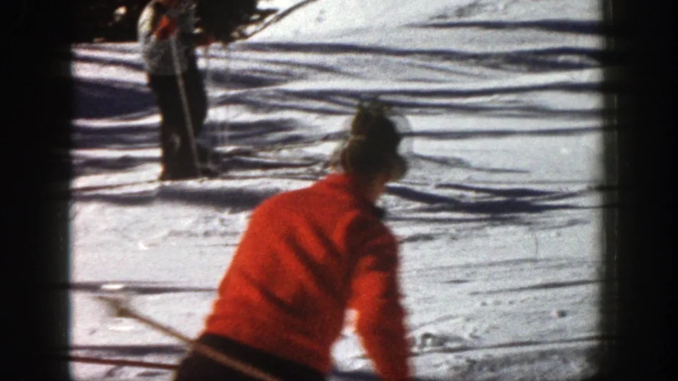 A skier in a red jacket moves downhill in Aspen, Colorado, with snow tracks, tree shadows, and another skier farther up the slope.