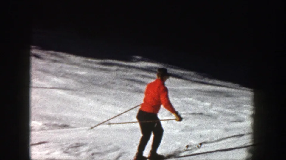 A red-jacketed skier continues downhill on the Aspen slope, framed against bright snow and a dark shadowed tree line.
