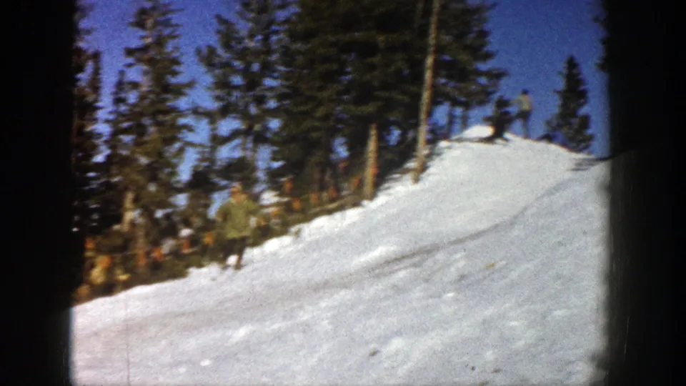 A distant skier in green outerwear descends a snowy Aspen slope in 1961, with tall evergreens and another skier near the ridge.