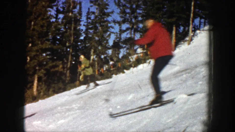 A red-clad skier passes near the camera on a snowy Aspen slope in 1961, with another skier and evergreens in the background.