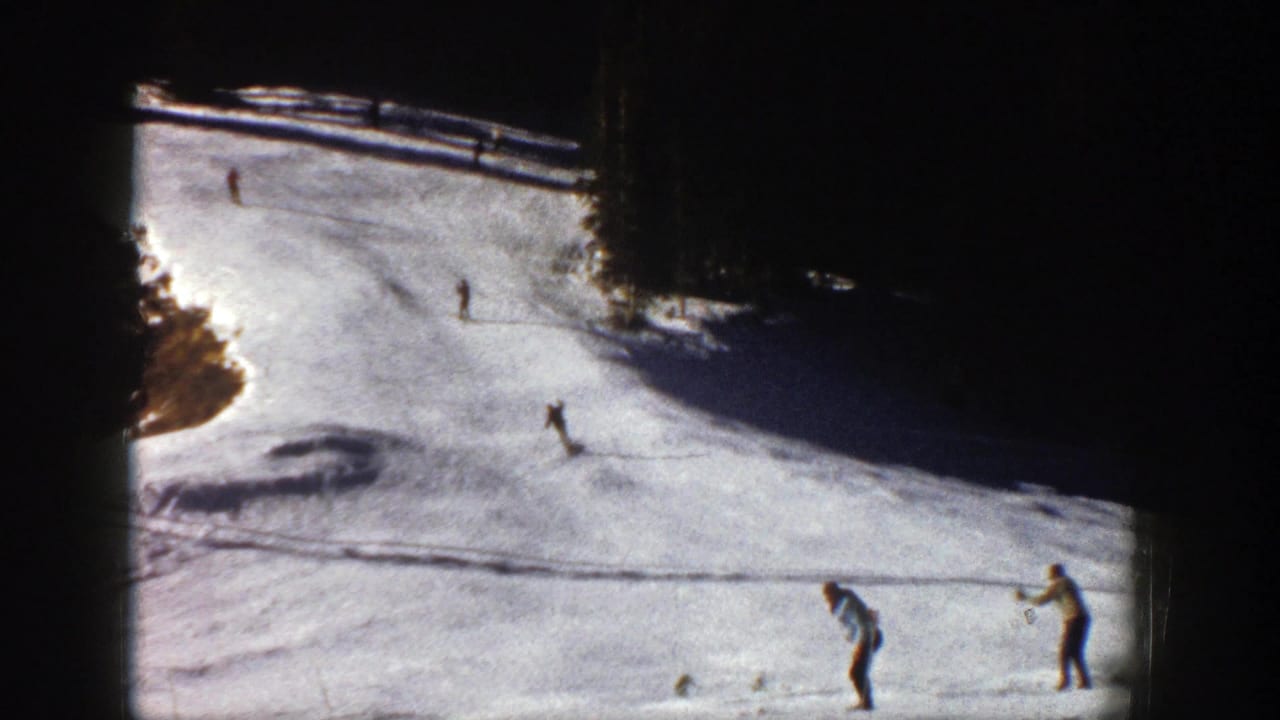 Skiers descend a snowy slope while two figures stand near the lower edge of the frame.