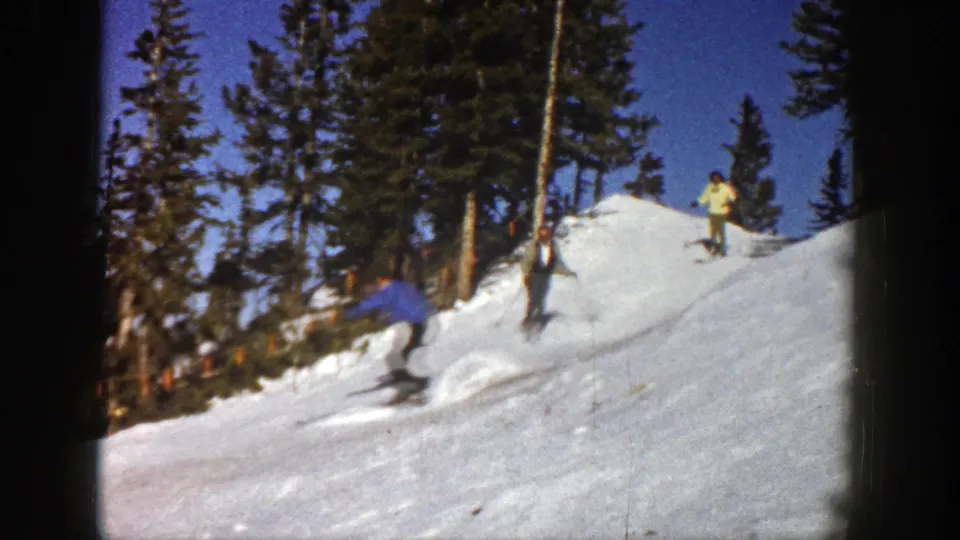 1961 Aspen frame showing a skier moving past the camera on a snowy slope, with pine trees and two standing skiers behind.