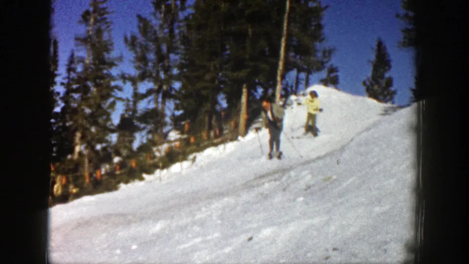 1961 Aspen 8mm frame of skiers paused on a bright snow slope, with tall pine trees and clear sky behind them.