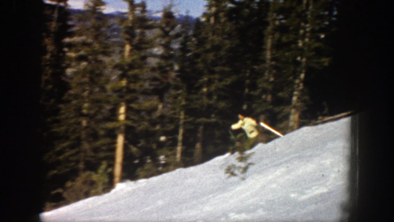 A skier descends a sunlit snowy slope in Aspen, Colorado, with evergreen trees and distant mountains in 1961 home-movie footage.