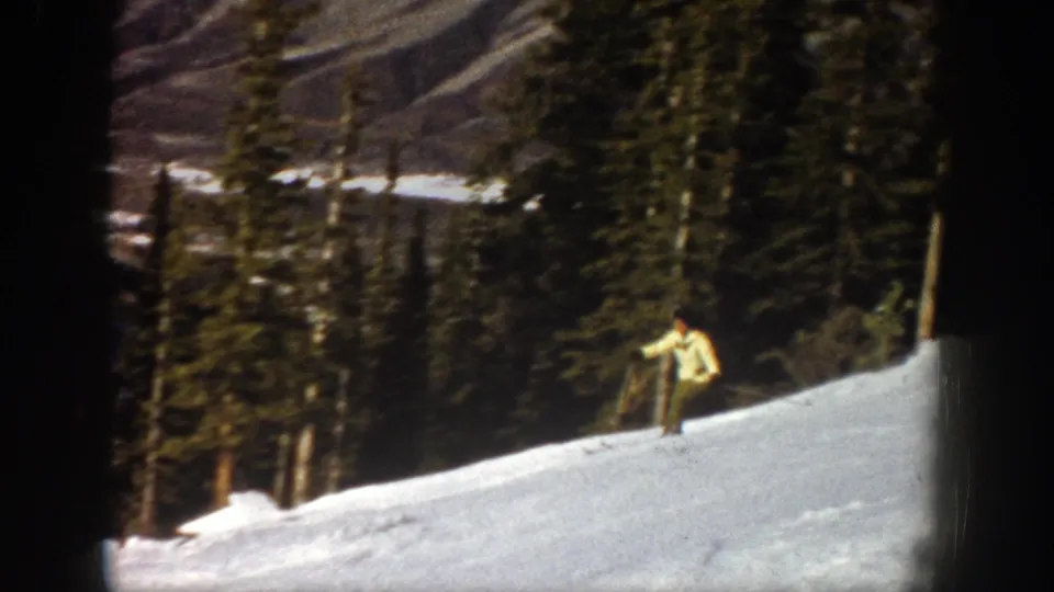 The skier continues across a bright Aspen snow slope, framed by dark evergreens and rocky mountain terrain in the distance.