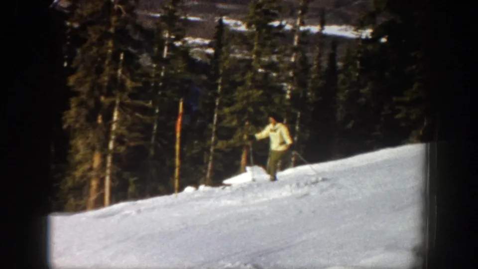 A distant skier in a light jacket moves lower along the Aspen slope, with tall evergreens filling much of the background.