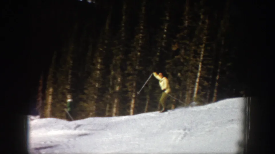 1961 Aspen home-movie frame showing a skier in a light jacket using poles near a dark evergreen forest.