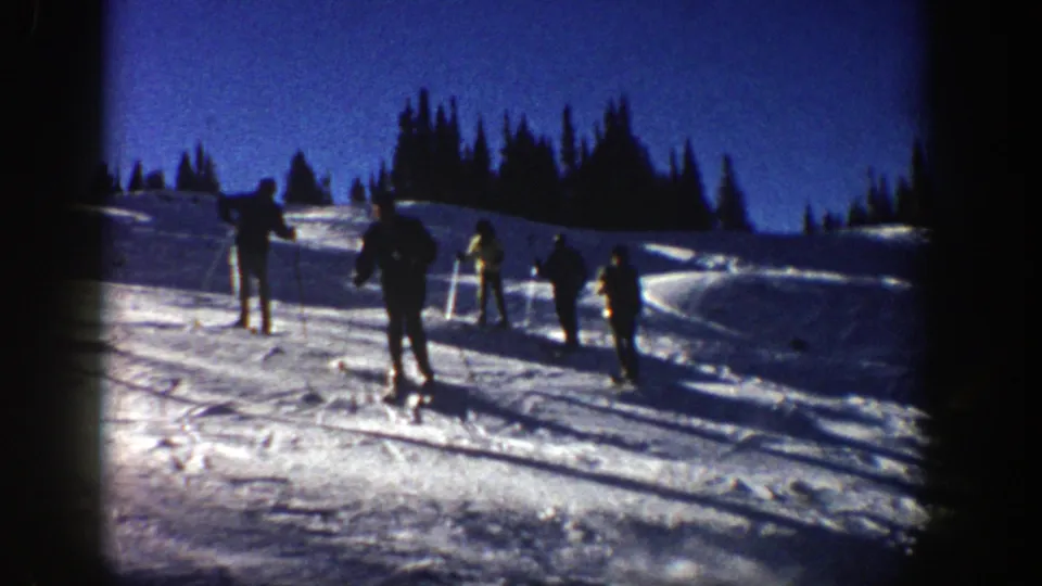 1961 Aspen home-movie frame looking across skiers moving away on tracked snow near evergreens.