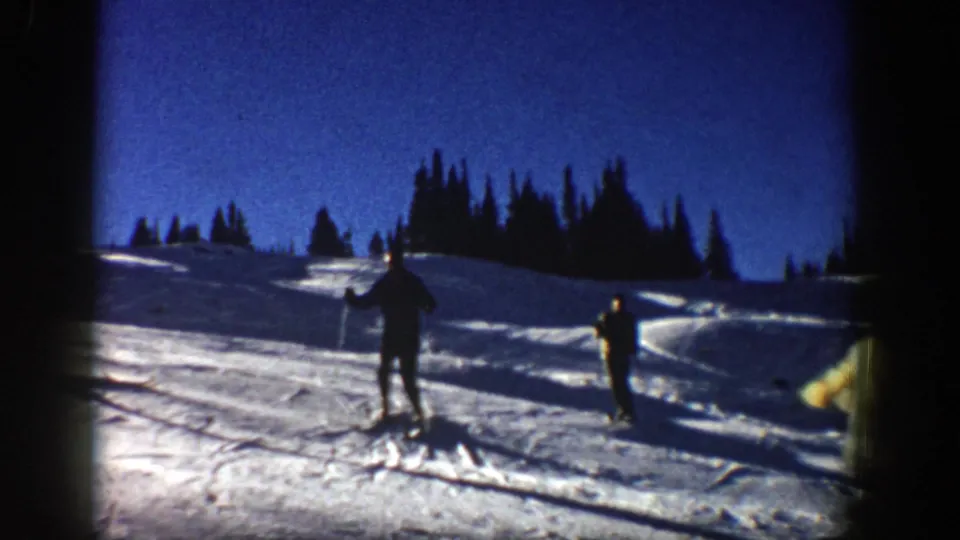 Two skiers continue down the Aspen slope beneath a blue sky, with dark evergreens lining the snowy ridge.