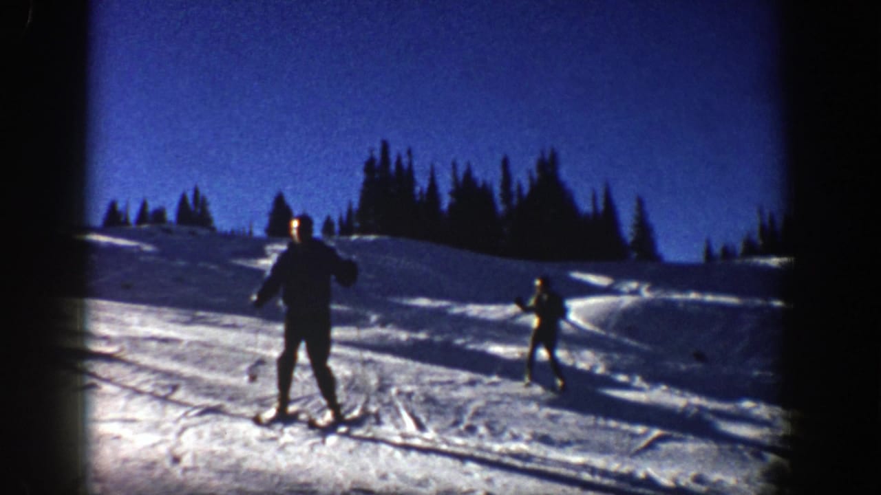 In 1961 Aspen home-movie footage, two adult skiers move across a bright snowy slope beneath a deep blue sky and dark pine ridgeline.