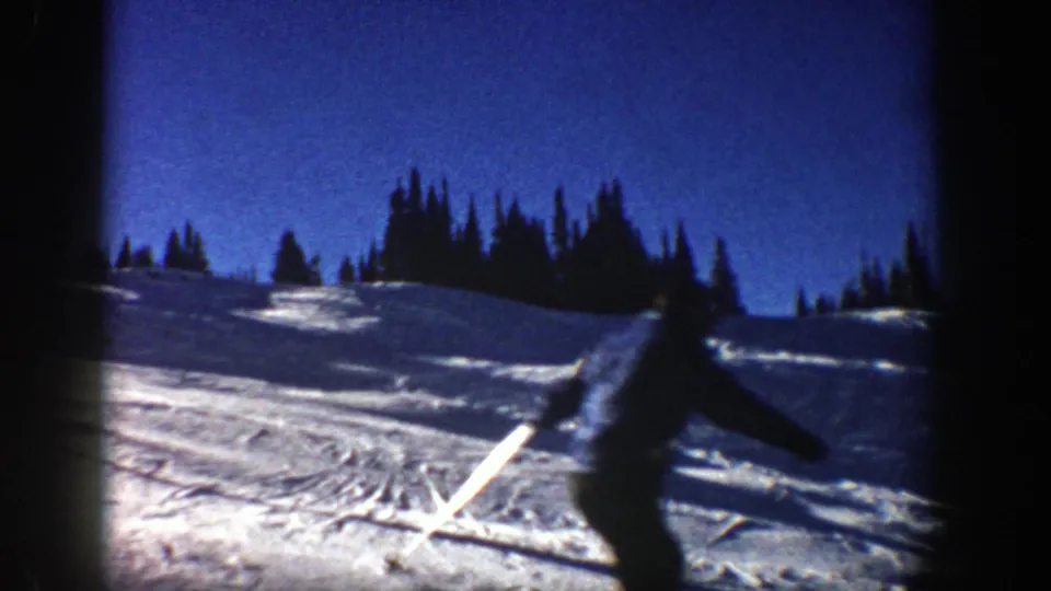 A silhouetted skier passes near the camera in 1961 Aspen 8mm footage, with snow, ski tracks, and pine trees visible behind.