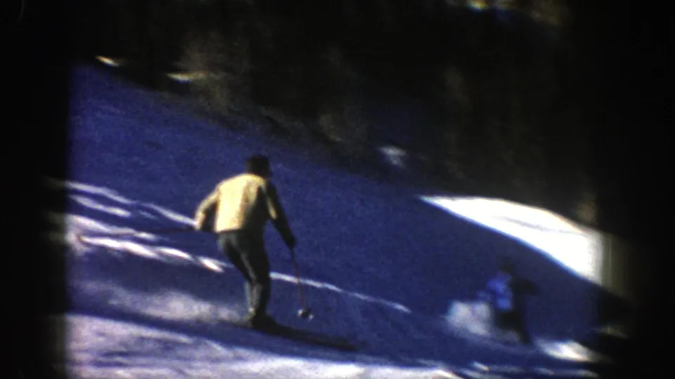 A skier in a light jacket descends a shaded Aspen slope, with dark trees, bright snow patches, and soft 8mm motion blur visible.