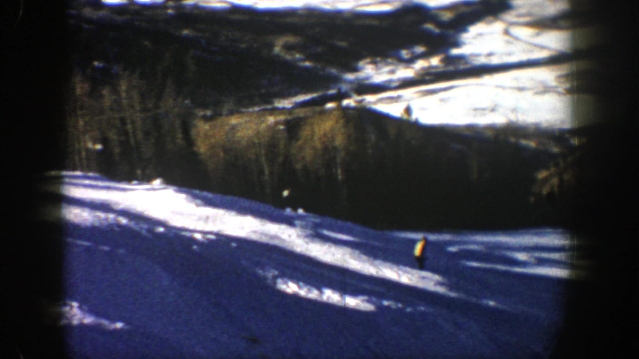 Snowy mountain ski slope with a distant skier in yellow and pine trees beyond.
