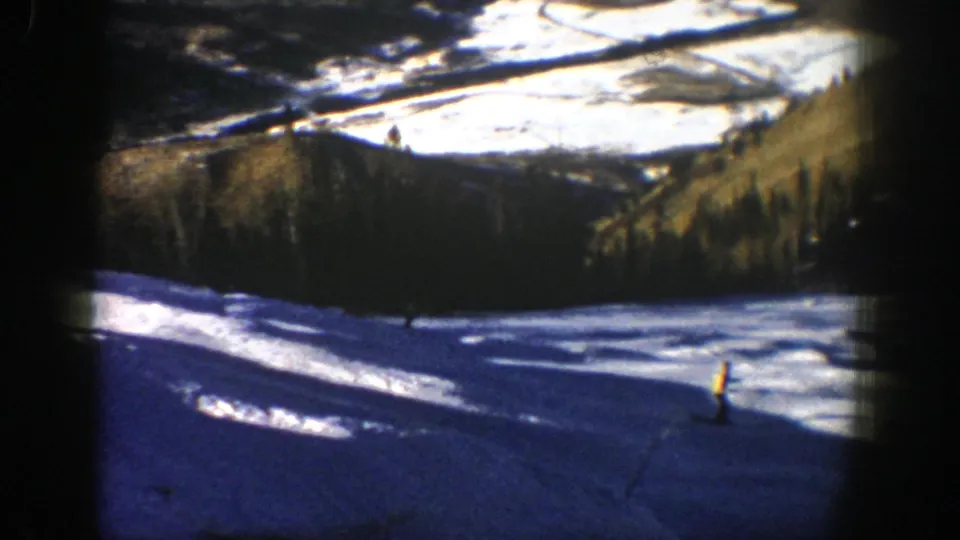 1961 Aspen frame with a small skier crossing a steep snow slope, framed by dark 8mm gate edges and pine-covered terrain.