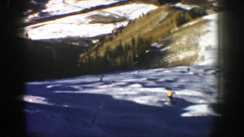 1961 Aspen home-movie frame showing multiple small skiers spread across a snow-covered run near pine-lined mountain slopes.