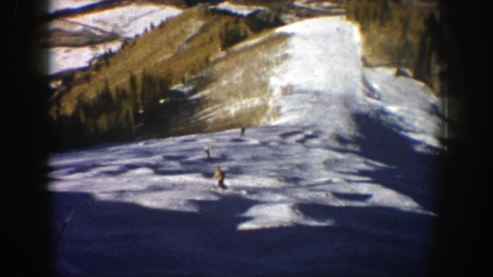 1961 Aspen frame with a yellow-jacketed skier descending a broad snow run, seen from a handheld 8mm home-movie viewpoint.