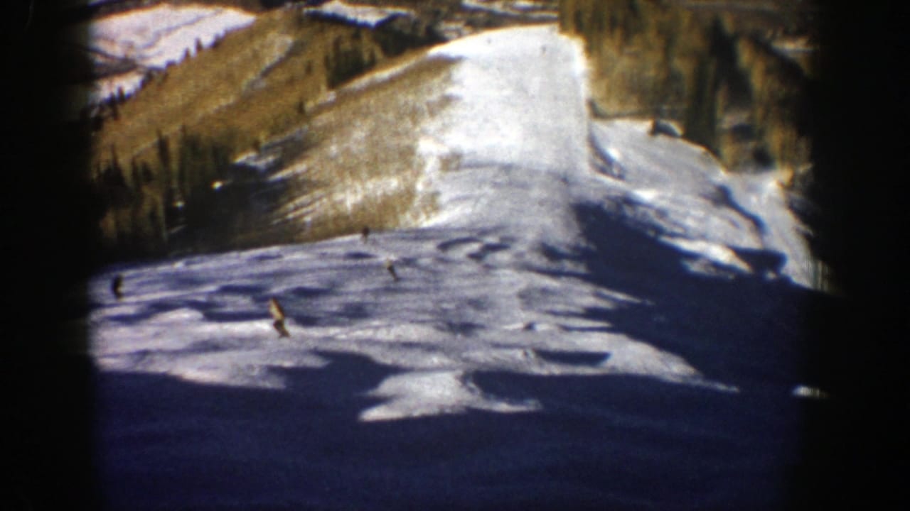 Snow-covered ski slope with small skiers and evergreen-lined mountain terrain.