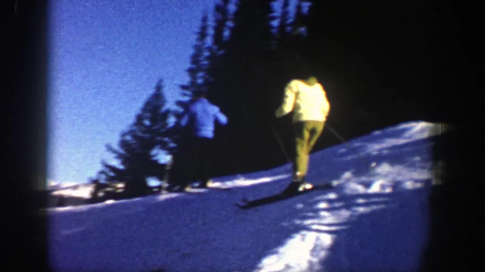 1961 Aspen home-movie frame showing two skiers on a snowy slope with dark evergreens behind them.