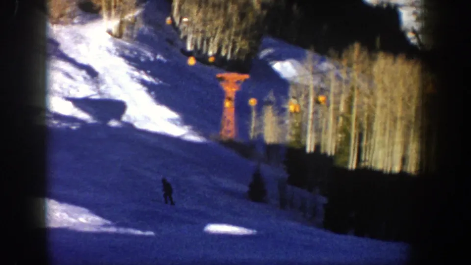 1961 Aspen ski-slope frame with a distant skier, blue-shadowed snow, orange lift structure, and winter trees lining the run.