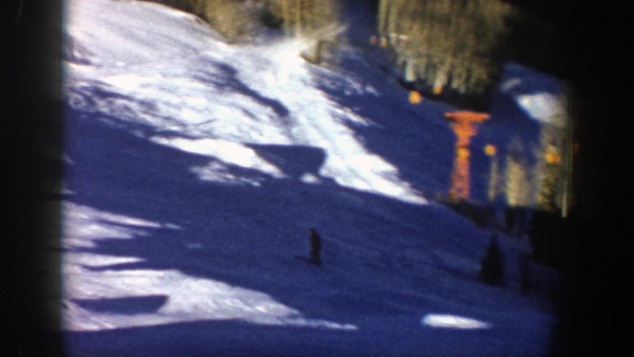 Snowy ski slope with a small skier, dark trees, strong shadows, and film gate edges.