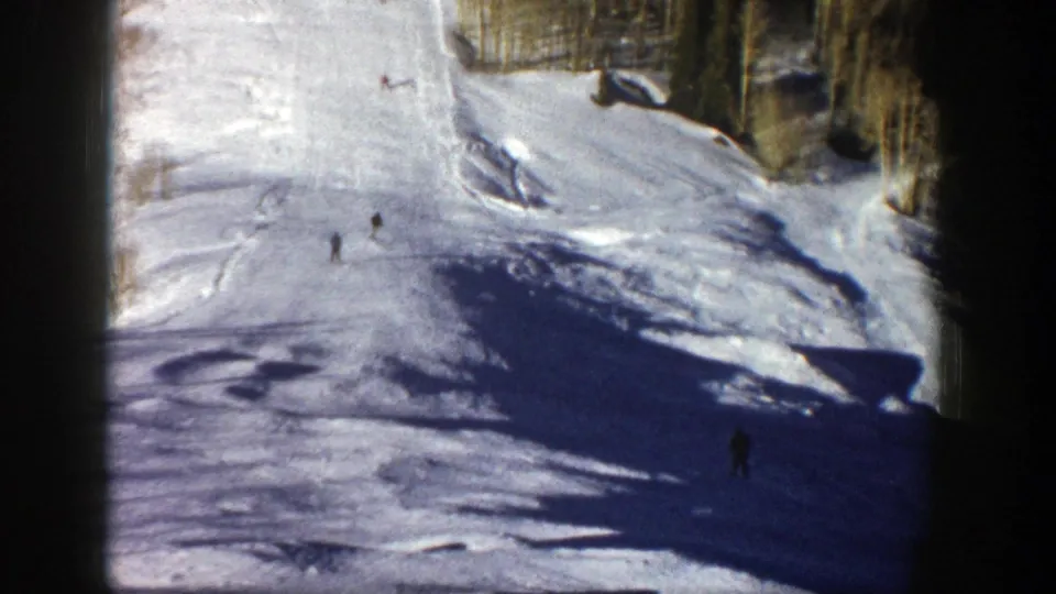 1961 Aspen, Colorado frame with several distant skiers on a bright open slope beside tall trees and patches of shadow.