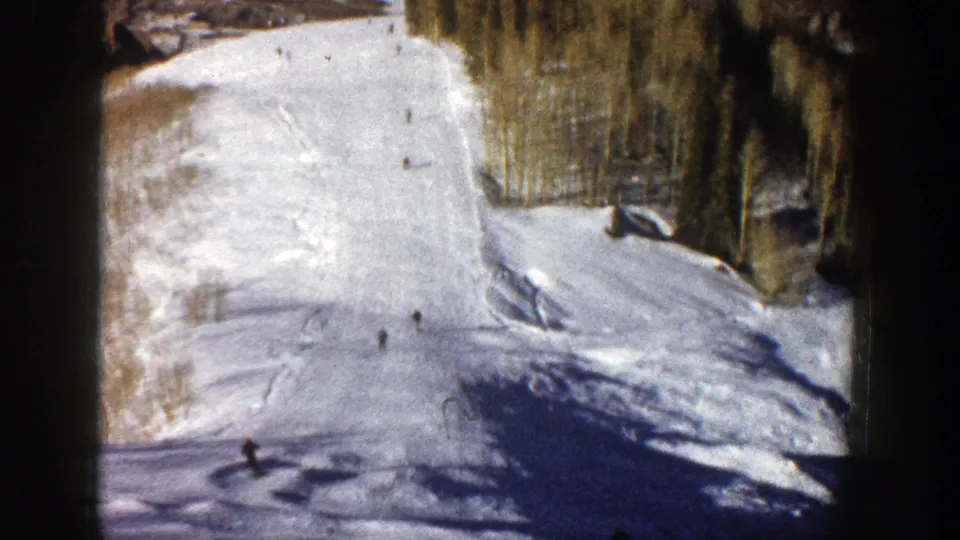 1961 Aspen, Colorado 8mm frame showing scattered skiers on a snowy run, with trees along the side and blue shadow across the foreground.