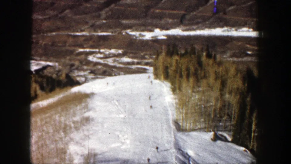 1961 Aspen, Colorado frame from an elevated viewpoint, showing tiny skiers on a snowy slope with trees and exposed terrain beyond.