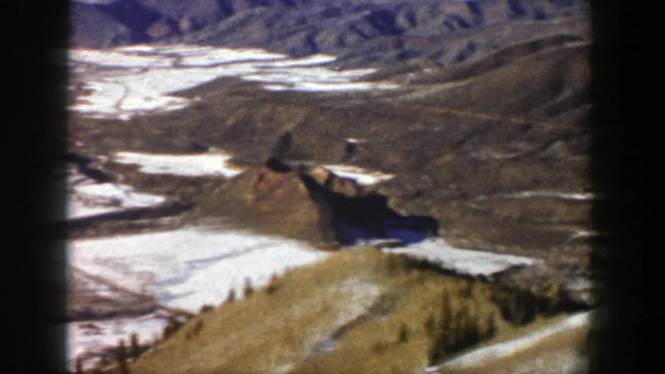 1961 Aspen, Colorado aerial home-movie frame with brown ridges, scattered melting snow, and winding terrain below a blue sky.