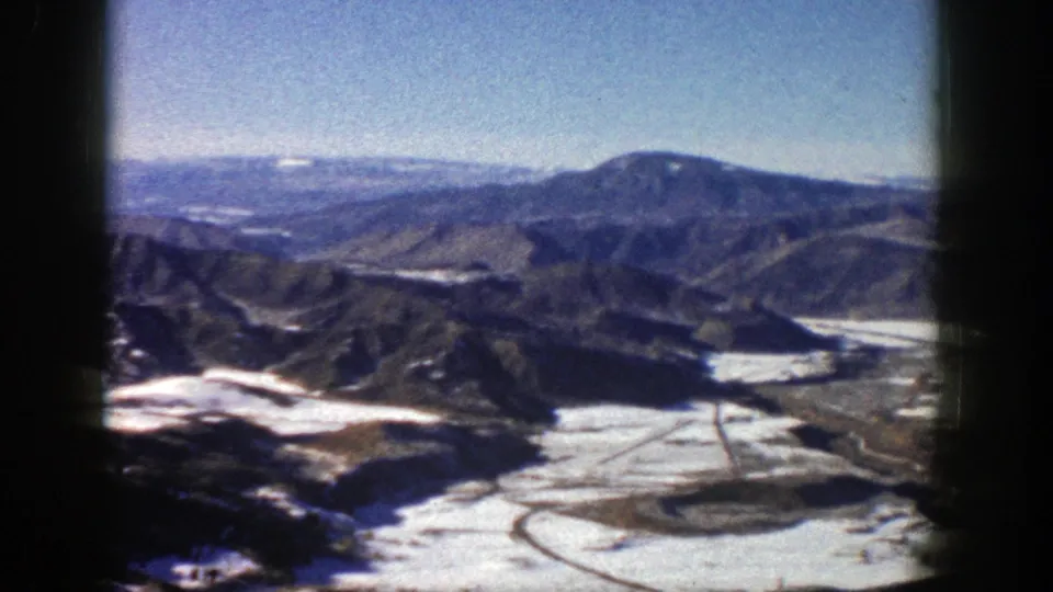 1961 Aspen, Colorado home-movie frame showing a high mountain valley, snow patches, blue sky, and layered distant ridges.