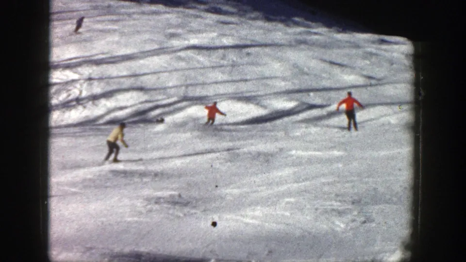 Multiple skiers cross a sunlit Aspen ski slope, with red and light-colored jackets visible against the snow.