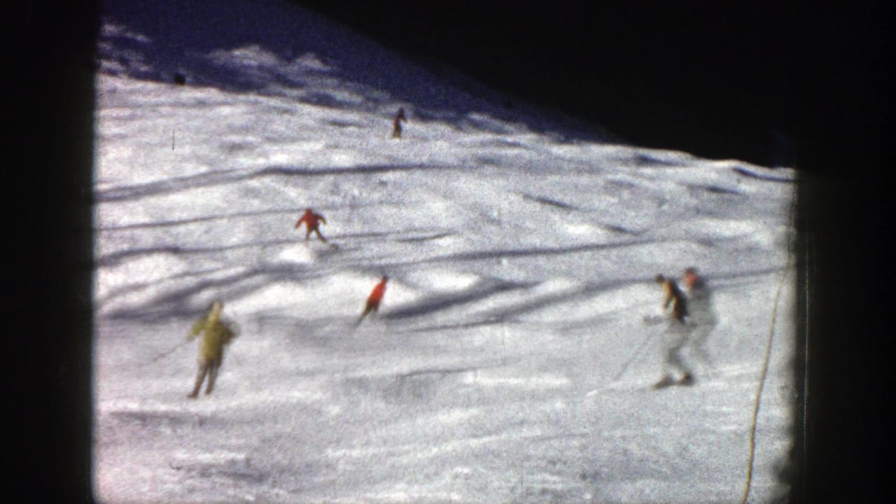 1961 Aspen home-movie frame showing several skiers spread across a bright snow slope with shaded terrain above.