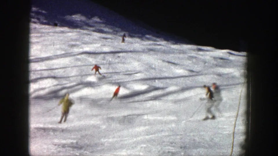 1961 Aspen home-movie frame showing several skiers spread across a bright snow slope with shaded terrain above.