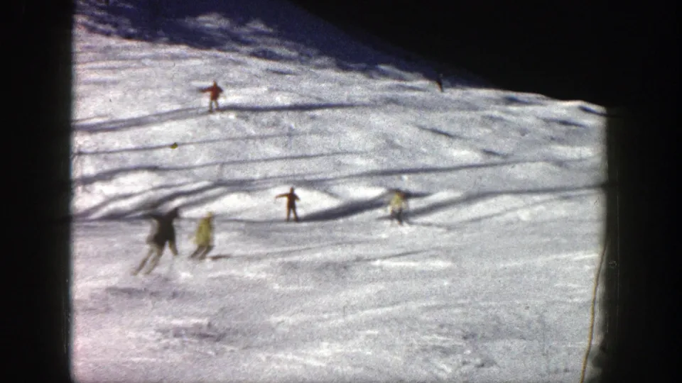 1961 Aspen ski-slope frame with distant skiers in red, yellow, and dark clothing moving through bright snow and shadow.