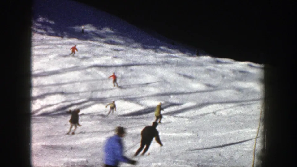 1961 Aspen home-movie still showing skiers at mixed distances, including closer dark and light figures near the bottom of the slope.