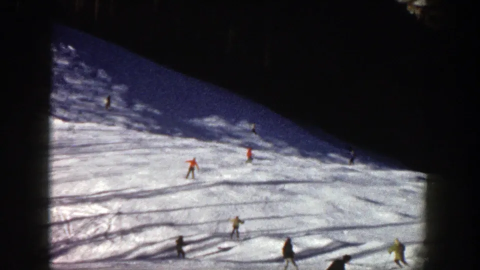 Wide 1961 Aspen ski-slope view with small skiers spaced across bright snow under a dark shaded upper hillside.