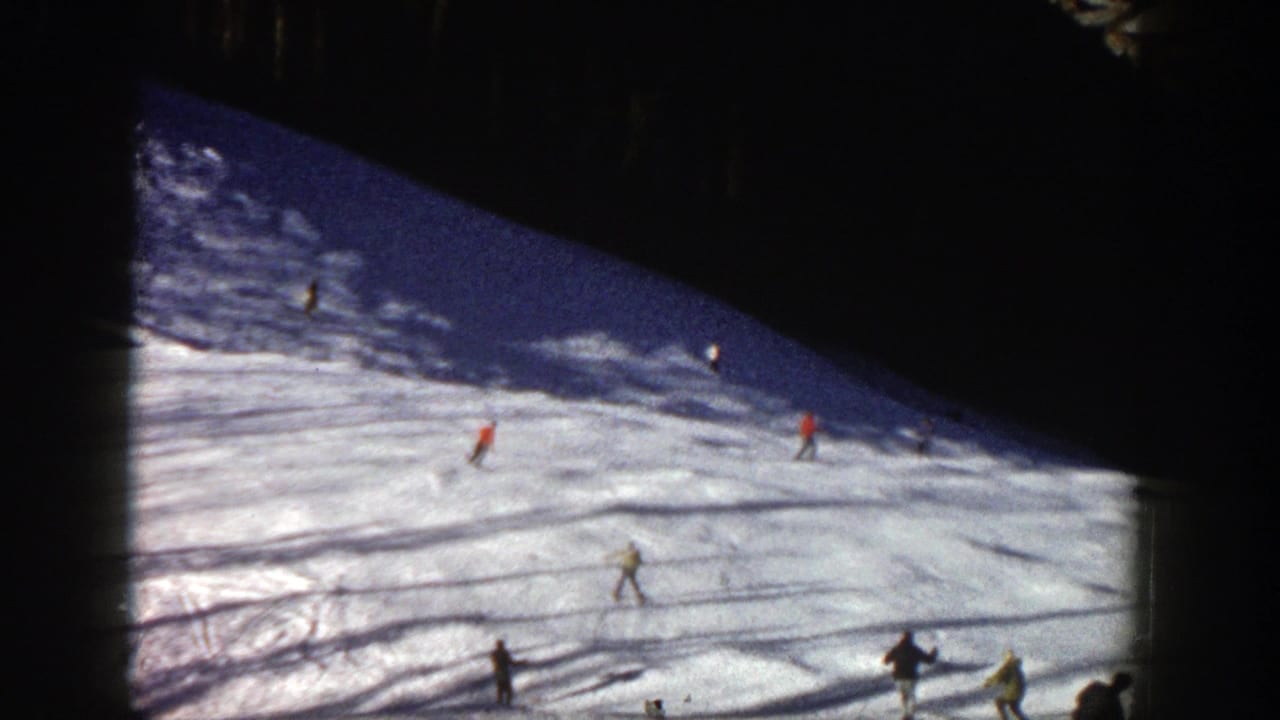 Skiers descend a sunlit snowy slope with dark film borders around the frame.