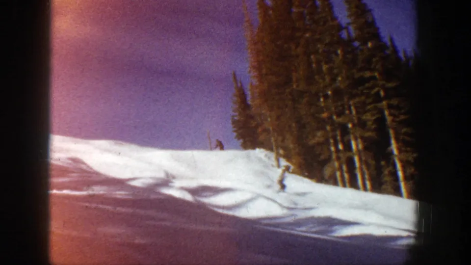 A skier descends a sunlit snowy slope in Aspen, Colorado, with dense evergreens and warm 8mm color shift visible.