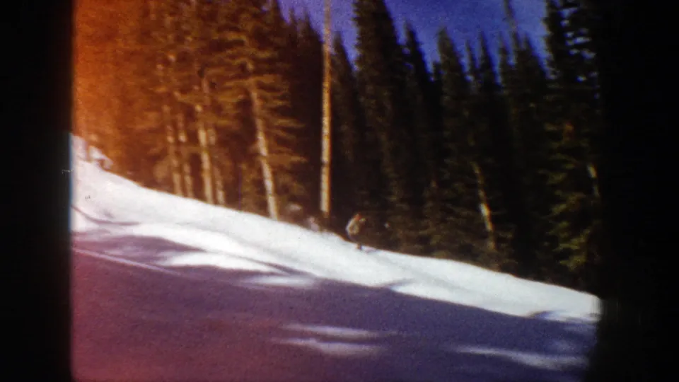 A small figure on skis crosses the lower part of a snowy Aspen slope, framed by tall evergreen trees and 8mm exposure variation.