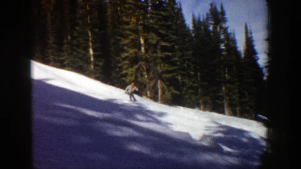 A skier is visible mid-descent on a snowy Aspen run in 1961, with evergreen forest, blue sky, and soft 8mm grain.