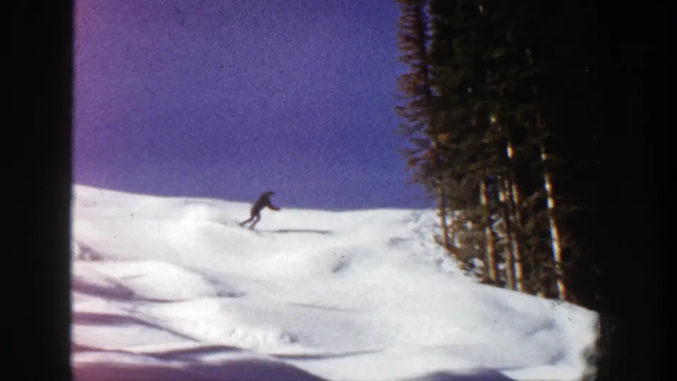 A lone skier in dark winter clothing descends a bright Aspen snow slope, with evergreen trees framing the run.