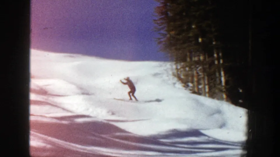Near the end of the 1961 Aspen clip, the skier continues downhill over tracked snow beside dark evergreens.