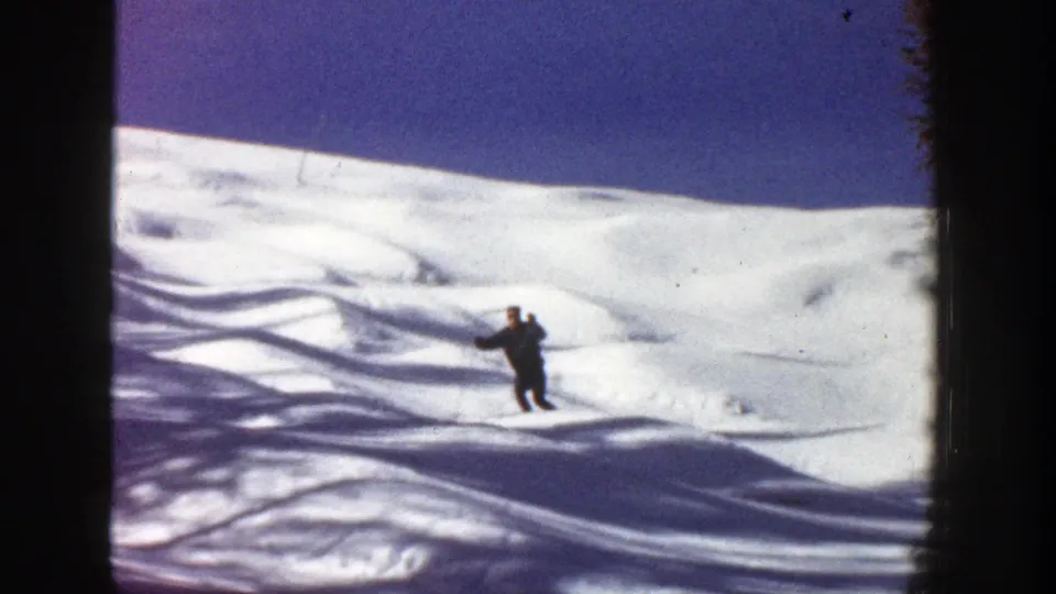 1961 Aspen, Colorado home-movie frame showing a distant skier on a white slope with purple-blue shadows and dark trees at the edge.