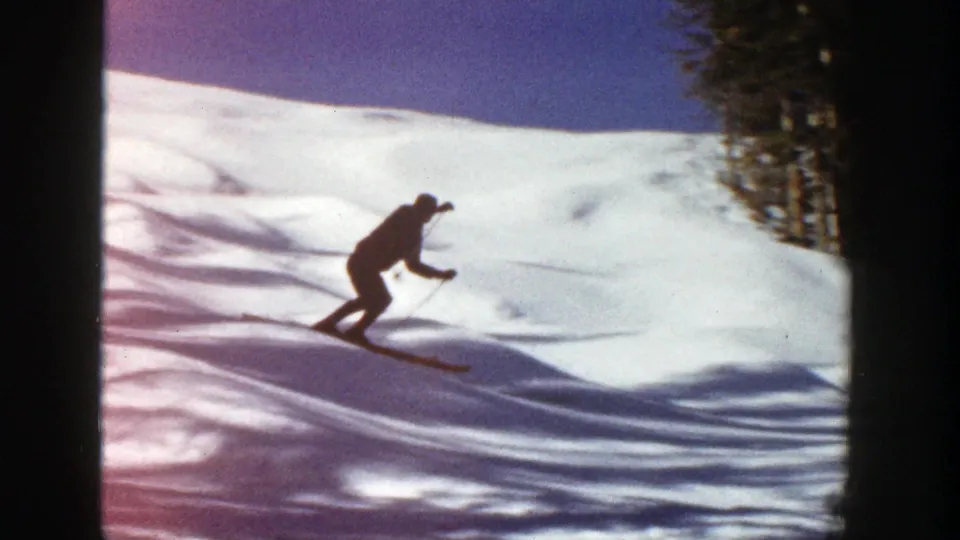 1961 Aspen, Colorado 8mm frame of a skier leaning into motion on a snow slope, with dark silhouette, tree line, and soft film grain.