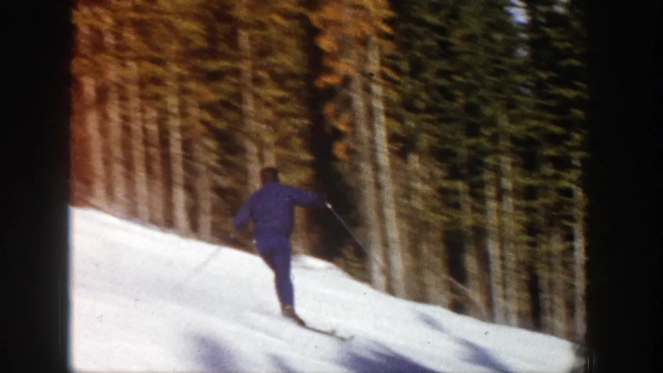 The skier moves away from the camera on a bright snow slope bordered by tall trees in 1961 Aspen footage.