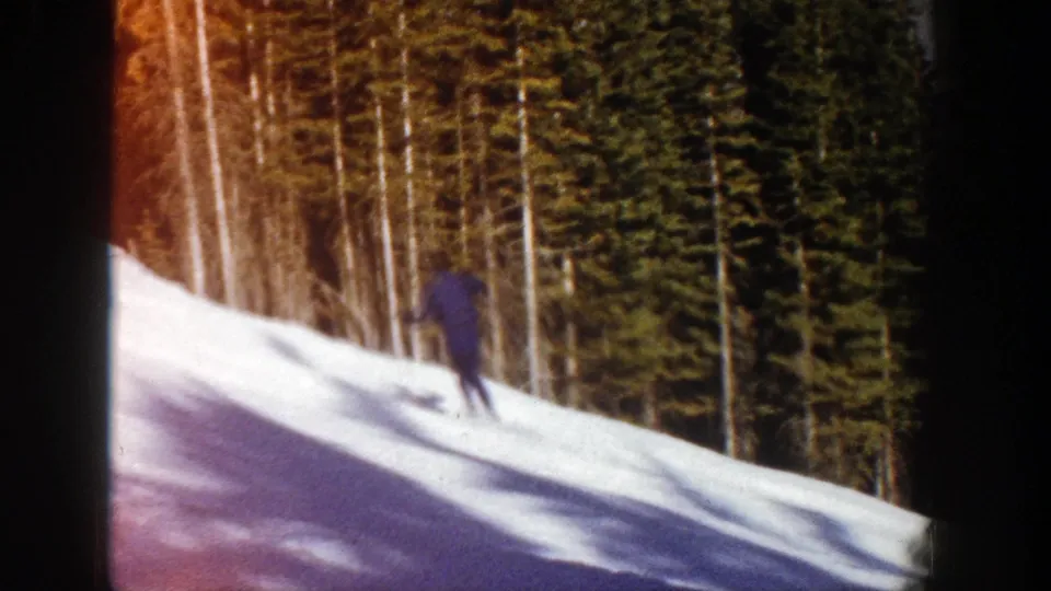 A distant skier crosses a sunlit snowy slope, with evergreen trees filling the background in Aspen, Colorado.