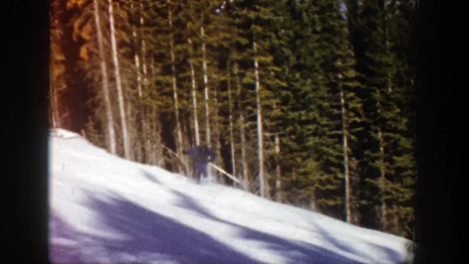 The skier appears small against the snowy slope and dense trees, continuing downhill in the 1961 home-movie clip.